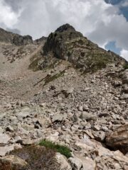 La falesia,in ombra, vista dal punto in cui si abbandona il sentiero. L'accesso avviene tramite l'evidente cengia erbosa di dx